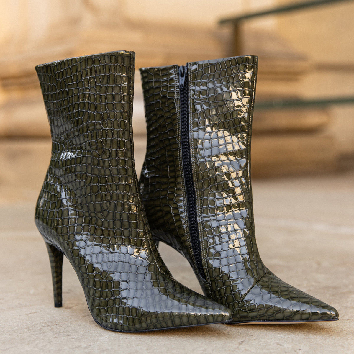 A pair of glossy olive green ankle boots with a crocodile-embossed texture, pointed toes, and slim stiletto heels, shown on a stone surface with a blurred background.