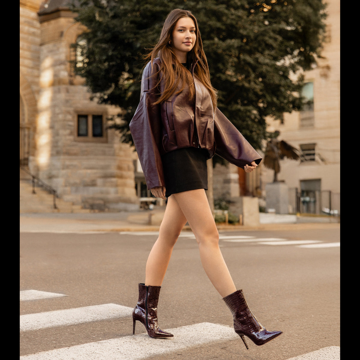 A woman walking across a city crosswalk wearing a shiny dark brown cropped jacket, a black mini skirt, and dark brown patent leather ankle boots with high heels, set against a background of a stone building and a large tree.
